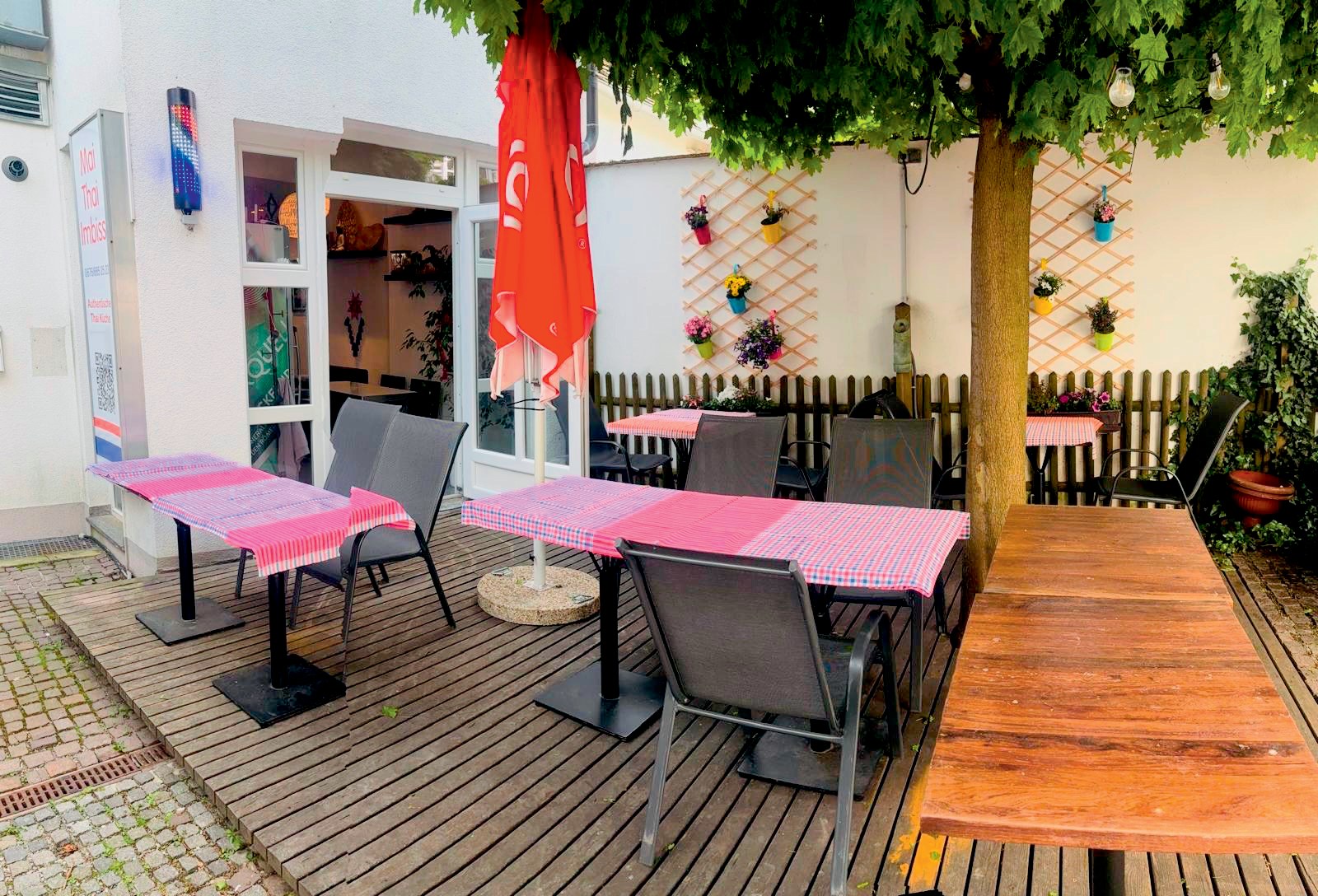 Gastgarten von Mai Thai: Tische mit karierten Tischtüchern auf einem Holzboden unter einem Baum, links der Eingang zum Lokal